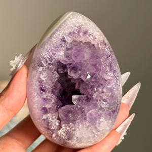 May include: An egg-shaped amethyst geode featuring a polished white outer shell and a deep, hollow center filled with jagged, translucent purple crystal points. The interior displays varying shades of violet, ranging from light lavender to deep purple.