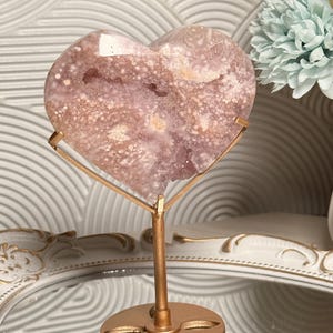 Puede incluir: Una geoda de cristal en forma de coraz&oacute;n, de color p&uacute;rpura claro, sobre un soporte de metal dorado. La geoda tiene un patr&oacute;n moteado con &aacute;reas blancas y p&uacute;rpuras m&aacute;s oscuras. El soporte tiene una base circular y un soporte vertical.