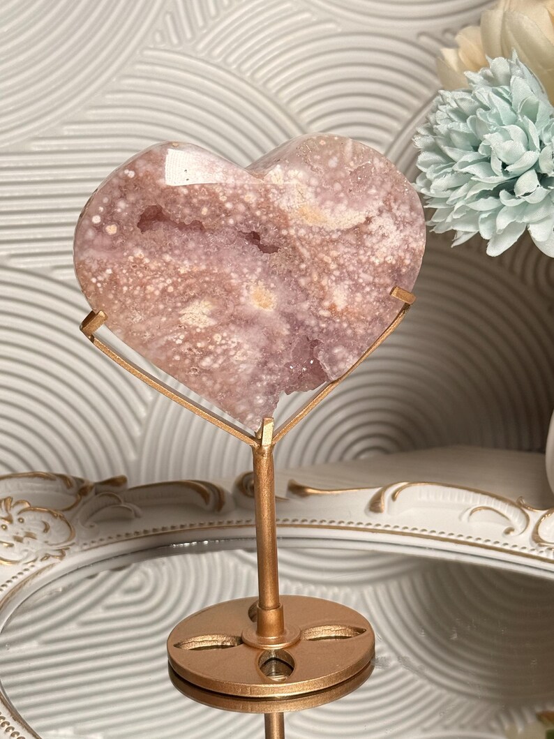 Puede incluir: Una geoda de cristal en forma de coraz&oacute;n, de color p&uacute;rpura claro, sobre un soporte de metal dorado. La geoda tiene un patr&oacute;n moteado con &aacute;reas blancas y p&uacute;rpuras m&aacute;s oscuras. El soporte tiene una base circular y un soporte vertical.