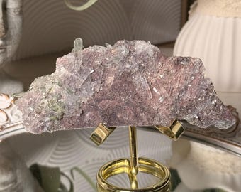 Può includere: Un esemplare minerale grezzo con una matrice piatta color marrone rossastro scuro, incastonata con numerosi piccoli cristalli di quarzo traslucidi a forma di ago. L'esemplare è sostenuto da un supporto espositivo in metallo dorato.