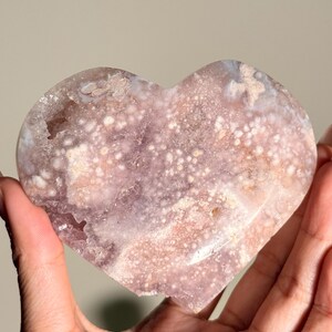 Puede incluir: Una piedra pulida en forma de coraz&oacute;n, con una superficie moteada y cristalina. La piedra presenta una mezcla de rosas suaves, p&uacute;rpuras y blancos. El coraz&oacute;n se muestra sobre un fondo neutro, resaltando su belleza natural.