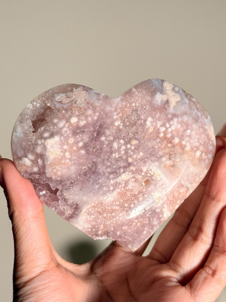 Puede incluir: Una piedra pulida en forma de coraz&oacute;n, con una superficie moteada y cristalina. La piedra presenta una mezcla de rosas suaves, p&uacute;rpuras y blancos. El coraz&oacute;n se muestra sobre un fondo neutro, resaltando su belleza natural.