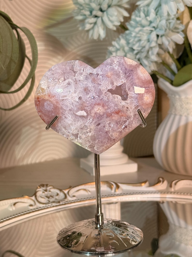 May include: A heart-shaped, polished crystal geode in shades of pink, purple, and white, mounted on a silver metal stand. The geode has a crystalline interior and is displayed against a blurred background with flowers.
