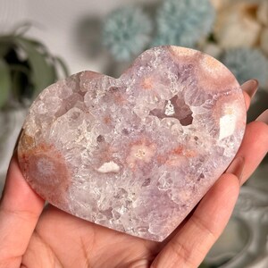K&ouml;nnte beinhalten: Eine herzf&ouml;rmige Amethyst-Geode mit kristallinem Innenraum und helllila und rosa Au&szlig;enbereich. Die Geode wird in einer Hand gehalten und zeigt ihre nat&uuml;rliche Sch&ouml;nheit und einzigartigen Muster. Die Geode ist ein dekorativer Gegenstand.