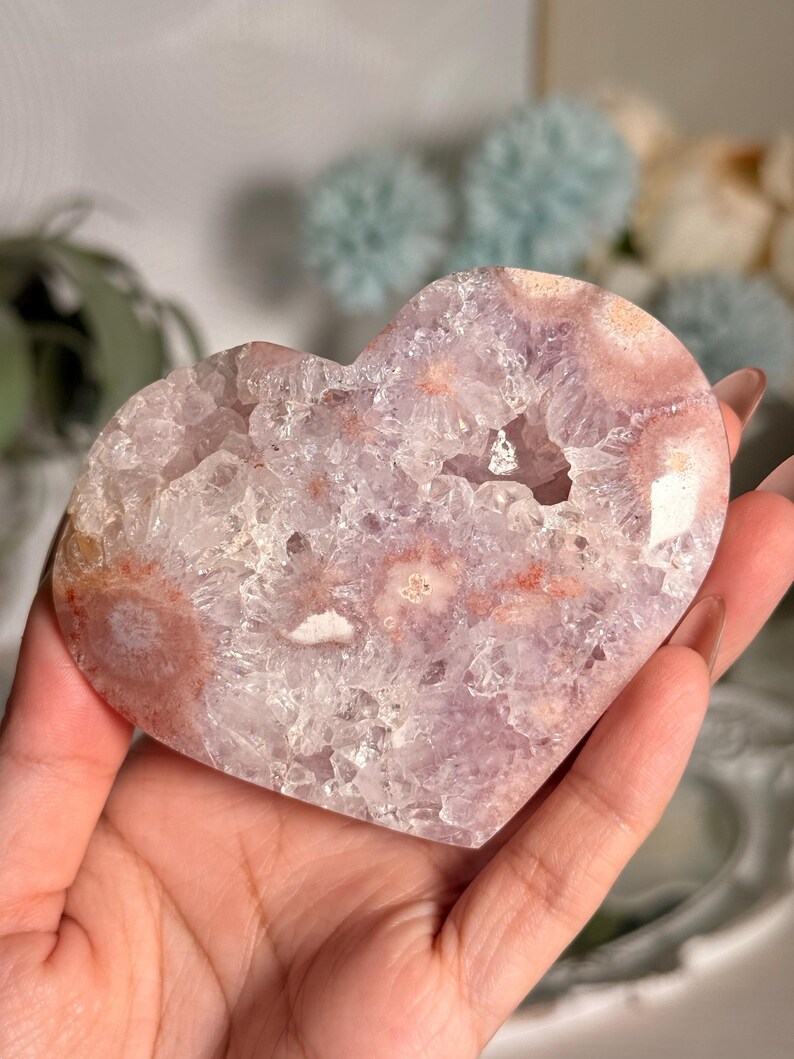 K&ouml;nnte beinhalten: Eine herzf&ouml;rmige Amethyst-Geode mit kristallinem Innenraum und helllila und rosa Au&szlig;enbereich. Die Geode wird in einer Hand gehalten und zeigt ihre nat&uuml;rliche Sch&ouml;nheit und einzigartigen Muster. Die Geode ist ein dekorativer Gegenstand.