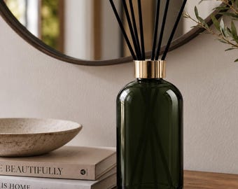 May include: A dark green glass reed diffuser bottle with a metallic gold collar and several thin black reeds extending from the top. The cylindrical bottle has a smooth, transparent finish and sits upright, showcasing a simple and functional design.