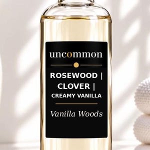 May include: A clear glass bottle with a silver cap containing a light amber liquid. The black rectangular label features the text uncommon, ROSEWOOD | CLOVER | CREAMY VANILLA, and Vanilla Woods in white and gold lettering on the front surface.