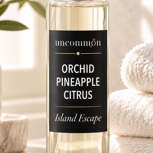 May include: A clear glass spray bottle with a silver cap containing a light yellow liquid. The black rectangular label features the text uncommon, ORCHID PINEAPPLE CITRUS, and Island Escape in white and gold lettering with a small gold icon.