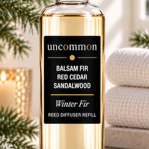 May include: A clear glass bottle of reed diffuser refill with a silver cap. The black rectangular label features the text uncommon, Balsam Fir, Red Cedar, Sandalwood, Winter Fir, and Reed Diffuser Refill in white and gold lettering.