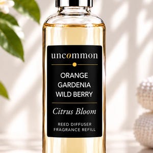 May include: A clear glass bottle filled with light amber liquid, featuring a black rectangular label that reads uncommon ORANGE GARDENIA WILD BERRY Citrus Bloom REED DIFFUSER FRAGRANCE REFILL. The bottle has a metallic silver cap.