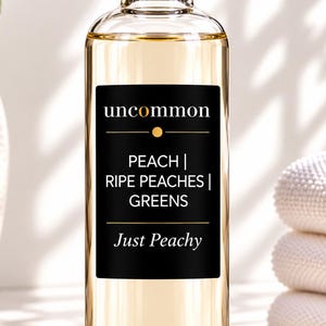 May include: A clear glass bottle filled with a light amber liquid, featuring a silver cap and a black rectangular label. The label displays the text uncommon, PEACH | RIPE PEACHES | GREENS, and Just Peachy in white and gold lettering.