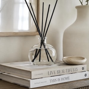 May include: A clear, round glass reed diffuser bottle filled with liquid and containing five thin black reeds. The diffuser sits atop two stacked books with the visible text "LIVE BEAUTIFULLY" and "THIS IS HOME" printed on their spines.
