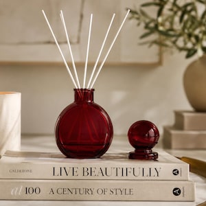 May include: A deep red glass diffuser bottle with a spherical body and a narrow neck, containing five thin white reed sticks. Beside the bottle sits a matching red glass spherical stopper with a small circular base, resting on two stacked books.