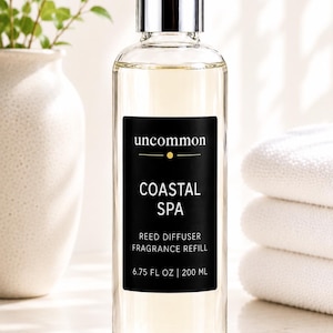 May include: A clear glass bottle filled with light-colored liquid, featuring a silver cap and a black rectangular label. The label reads uncommon COASTAL SPA REED DIFFUSER FRAGRANCE REFILL 6.75 FL OZ | 200 ML in white and gold text.