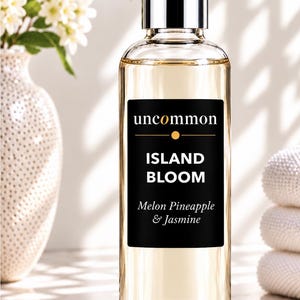May include: A clear glass bottle filled with light amber liquid, featuring a silver cap and a black rectangular label. The label displays the text uncommon, ISLAND BLOOM, and Melon Pineapple & Jasmine in white and orange lettering.