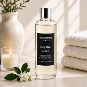 May include: A clear glass bottle of reed diffuser fragrance refill with a silver cap and a black label reading uncommon CABANA COVE REED DIFFUSER FRAGRANCE REFILL 6.75 FL OZ | 200 ML. The liquid inside is light yellow and the bottle is cylindrical.