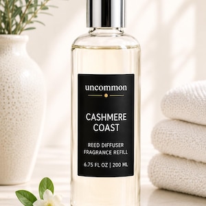 May include: A clear glass bottle of reed diffuser fragrance refill with a silver cap and a black label. The label reads uncommon CASHMERE COAST REED DIFFUSER FRAGRANCE REFILL 6.75 FL OZ | 200 ML. The bottle contains a light-colored liquid.