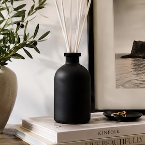 May include: A matte black glass reed diffuser bottle with a narrow neck, containing several thin white reed sticks. The bottle sits atop a stack of two books, one labeled KINFOLK and the other labeled LIVE BEAUTIFULLY by CALDERONE.