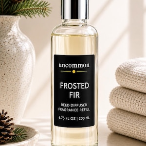 May include: A clear glass bottle of Uncommon Frosted Fir reed diffuser fragrance refill with a silver cap. The black label features white text reading "uncommon", "FROSTED FIR", "REED DIFFUSER FRAGRANCE REFILL", and "6.75 FL OZ | 200 ML".