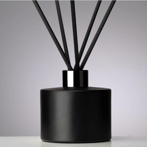 May include: A black diffuser bottle with a silver collar and several black reeds. The cylindrical bottle has a matte finish. The reeds are arranged to fan out from the top of the bottle. Home fragrance product.