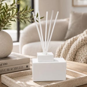 May include: A white rectangular ceramic reed diffuser vessel with a matching square cap and five slender white diffuser reeds inserted into the top. The diffuser has a smooth matte finish and sits on a light-colored textured stone tray.