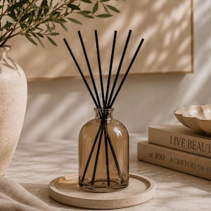 May include: A translucent amber glass reed diffuser bottle with a short neck and a rounded base, containing six thin black reed sticks arranged in a fan shape, resting on a circular beige stone tray.
