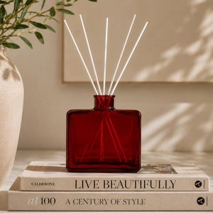 May include: A square, deep red glass reed diffuser bottle with five thin white reeds inserted into the neck. The bottle sits atop two stacked books, with the top book titled "LIVE BEAUTIFULLY" and the bottom book titled "at 100 A CENTURY OF STYLE."