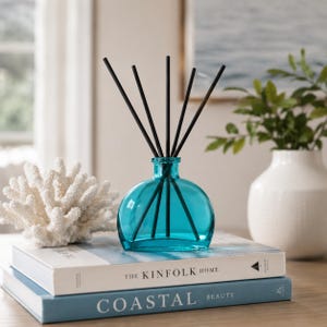 May include: A teal glass reed diffuser bottle with five black reeds, positioned on top of two stacked books titled The Kinfolk Home and Coastal Beauty, accompanied by a white decorative coral piece and a white ceramic vase with green foliage.