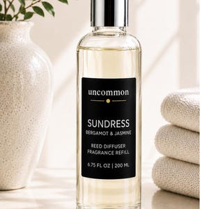 May include: A clear glass bottle of reed diffuser fragrance refill with a silver cap. The black label reads uncommon SUNDRESS BERGAMOT & JASMINE REED DIFFUSER FRAGRANCE REFILL 6.75 FL OZ | 200 ML. The liquid inside is a pale yellow color.