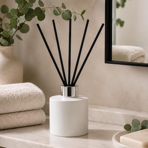 May include: A white cylindrical reed diffuser with a silver-toned metal collar and five thin black reeds extending upwards. The diffuser has a smooth, matte finish and a simple, clean design, standing upright as a functional home fragrance accessory.