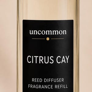 May include: A clear glass bottle filled with light yellow liquid, topped with a silver cap. A black rectangular label features the white text: uncommon, CITRUS CAY, REED DIFFUSER FRAGRANCE REFILL, 6.75 FL OZ | 200 ML. The bottle has a cylindrical shape.