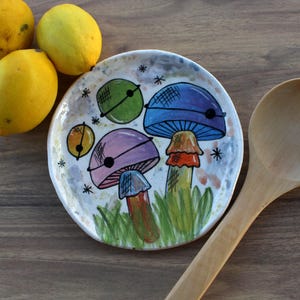 May include: A hand-painted ceramic plate featuring mushroom illustrations in blue, purple, and orange, with a wooden spoon and three lemons. The items are arranged on a wooden surface, creating a rustic aesthetic.