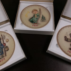 May include: Three decorative plates in white boxes. Each plate features a different illustration of a child in a winter scene, with the year printed below. The plates are in a cream color with a brown border.