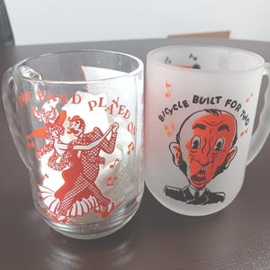 May include: Two glass mugs with vintage-style designs. One mug is clear with a red illustration of dancers and the text "THE BAND PLAYED ON." The other is frosted with a cartoon face and the text "BICYCLE BUILT FOR TWO."