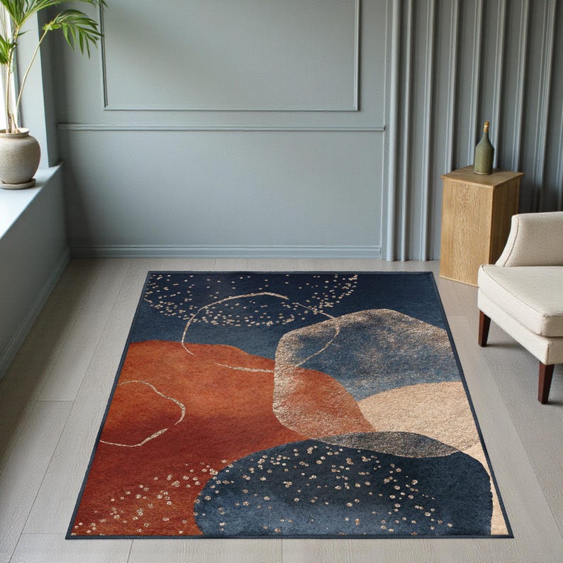 May include: A rectangular area rug with an abstract design. The rug features a dark blue background with large, overlapping shapes in shades of rust, beige, and light blue. White speckles and curved lines add detail to the design. The rug is placed on a light wood floor.
