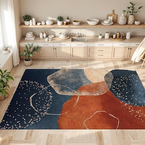 May include: A rectangular area rug with an abstract design. The rug features a dark blue background with rust, beige, and gold-colored shapes. The rug is placed on a light-colored wooden floor in a room with neutral-toned walls and cabinets.