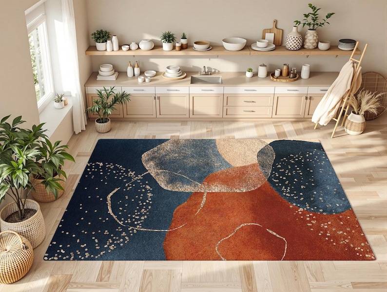 May include: A rectangular area rug with an abstract design. The rug features a dark blue background with rust, beige, and gold-colored shapes. The rug is placed on a light-colored wooden floor in a room with neutral-toned walls and cabinets.