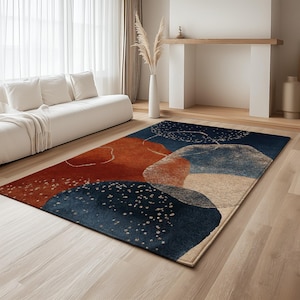 May include: A rectangular area rug with an abstract design. The rug features a dark blue background with rust, beige, and light blue organic shapes. White speckles and thin white lines add detail to the design. The rug is placed on a light wood floor.