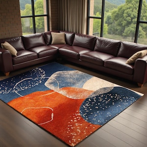 May include: A modern area rug with an abstract design in shades of navy blue, orange, and white. The rug is placed on a hardwood floor in front of a burgundy leather sectional sofa. The rug's design features organic shapes and scattered white dots.
