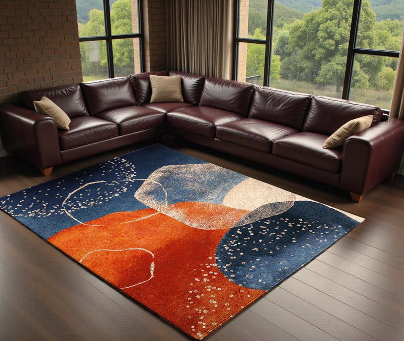 May include: A modern area rug with an abstract design in shades of navy blue, orange, and white. The rug is placed on a hardwood floor in front of a burgundy leather sectional sofa. The rug's design features organic shapes and scattered white dots.