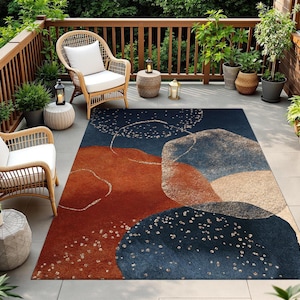 May include: An outdoor rug with an abstract design in shades of navy blue, rust, and beige. The rug features a pattern of overlapping shapes and scattered white dots. The rug is placed on a patio with wicker chairs and potted plants.