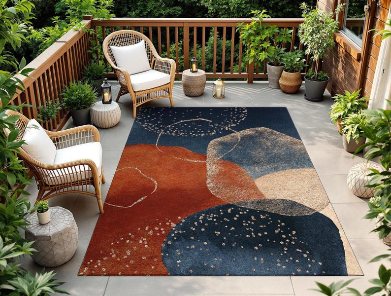May include: An outdoor rug with an abstract design in shades of navy blue, rust, and beige. The rug features a pattern of overlapping shapes and scattered white dots. The rug is placed on a patio with wicker chairs and potted plants.