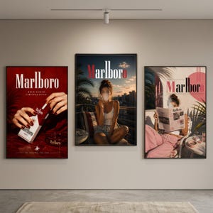 May include: Three framed wall art prints featuring Marlboro branding. The left print shows hands holding a cigarette pack on a red background. The centre print depicts a person on a balcony at sunset. The right print shows a person reading a newspaper.