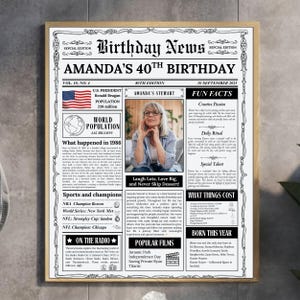 May include: A framed newspaper-style poster titled Birthday News Amanda's 40th Birthday. The black and white print features a central photograph of a person, a US flag, and columns of text detailing fun facts, world population, and 1986 events.