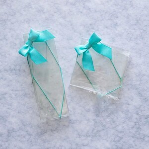 May include: Two clear cellophane gift boxes tied with turquoise satin bows. The boxes are decorated with turquoise thread and the text "To The Occasional Cookier". One box is rectangular, the other is square.