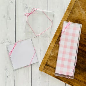 May include: Three clear, square boxes tied with pink string. One box contains a pink and white gingham pattern, while the others hold white paper. The boxes are on a white wooden surface and a wooden board.