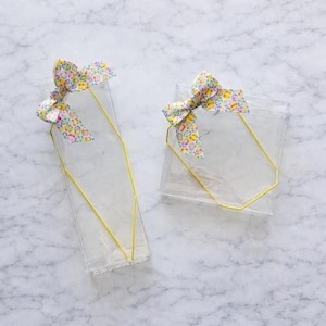 May include: Two clear cellophane bags with yellow wire handles and colorful patterned bows. The bags are rectangular and have a simple, elegant design. The bows feature a mix of pastel colors and patterns.