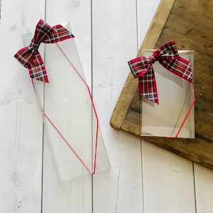 May include: Two clear plastic gift boxes with red ribbon bows. The boxes are on a white wooden surface. The bows are red and black plaid.