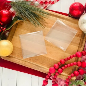 May include: Two clear plastic square gift boxes on a wooden cutting board surrounded by red and white checkered fabric, red Christmas ornaments, a gold ornament, a white ornament, and a strand of red wooden beads.
