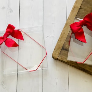 May include: Two clear, hexagonal gift boxes with bright red bows. Each box has a red string outline. One box is on a white wooden surface, and the other is on a wooden surface.